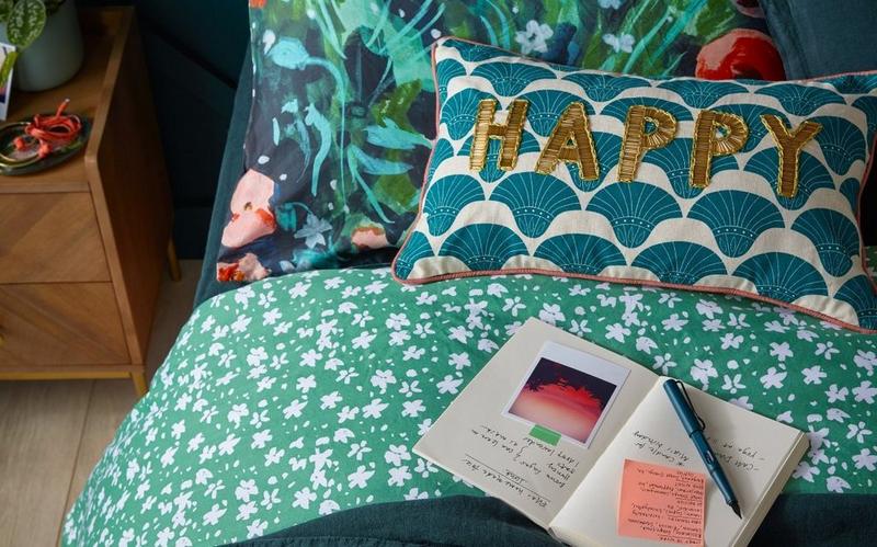 close up of green floural bedding including a cushion with 'happy' in gold embroidary and an open journal on the bed